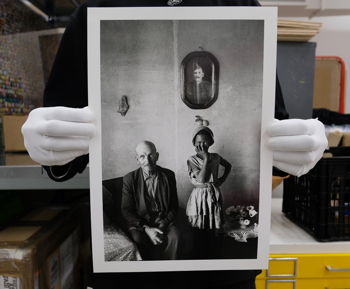 Process photograph from the ‘Picture Theory’ exhibition in A4’s Gallery. Hands wearing archive gloves hold a printed copy of David Goldblatt’s photograph ‘A plot-holder with the daughter of his servant, Wheatlands, Randfontein’. The plot-holder sits on the left, with his servant’s daughter on the right.
