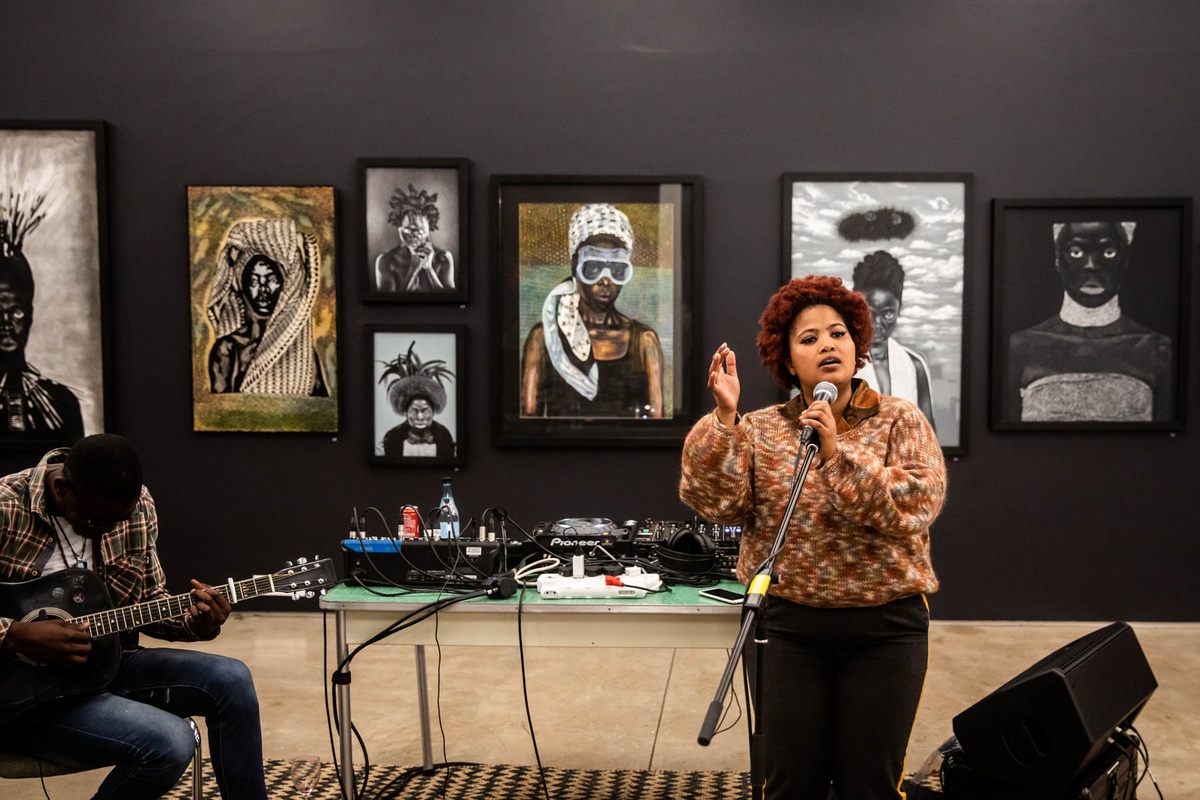 Event photograph from ‘Finissage: Ikhono LaseNatali’ in A4’s Gallery. At the front, vocalist Michelle Fani with a microphone stand, with a seated guitarist on the left, and a table laden with audio equipment behind them. At the back, artworks from the ‘Ikhono LaseNatali’ exhibition are mounted on the wall.
