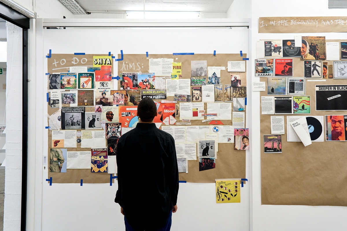 Process photograph from the Black Plastic residency in A4 Arts Foundation. At the back, panels of brown paper are taped to a white wall, collaged with printouts of vinyl record covers and typed text. The panel headings read "To do" and "Aims".
