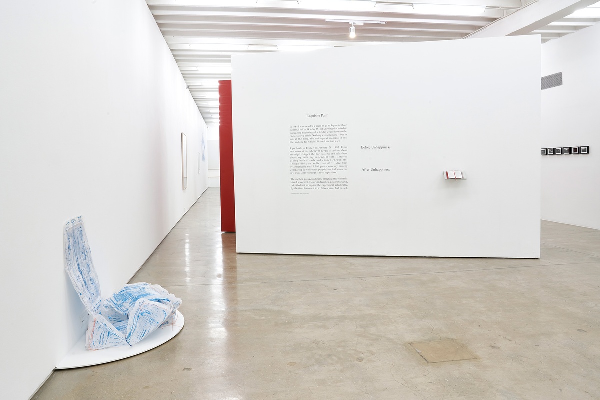 Installation photograph of the You to Me, Me to You exhibition. On the left, Thato Makatu’s sculptural butcher-paper work ‘home is…’ sits on a white semi-circular platform attached to the base of a white gallery wall. At the back, Sophie Calle’s ‘Exquisite Pain’ is arranged on a white moveable gallery wall, with a vinyl wall text on the left and a book on a wall-mounted shelf on the right.
