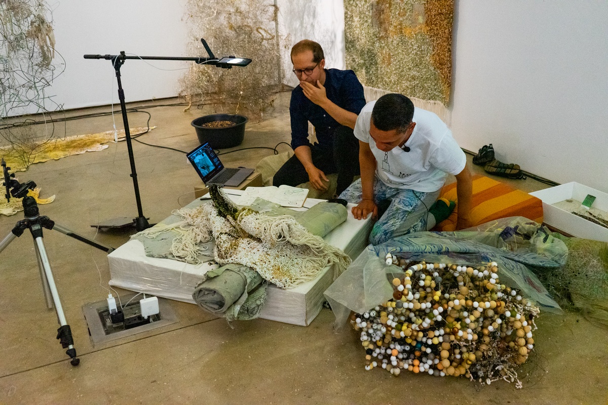 Event photograph from ‘Knots’, a conversation between Igshaan Adams and Josh Ginsburg, marking the close of ‘Open Production’, Adams’ hybrid studio/exhibition in A4’s Gallery. On the right, Adams and Ginsburg examine one of Adams’ woven tapestries under a recording device mounted on a stand.
