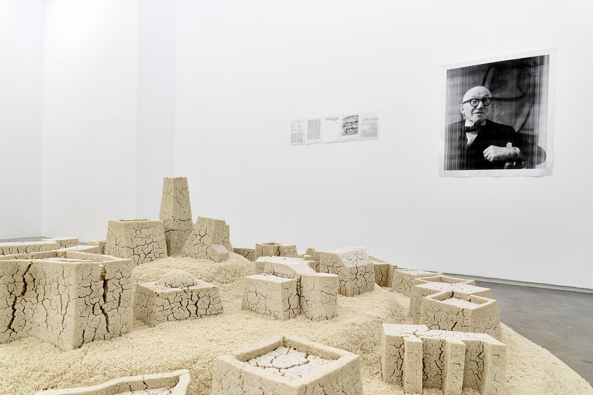 Installation photograph from The Future is Behind Us exhibition in A4’s Gallery. At the front, Kader Attia’s couscous sculpture from ‘Untitled (Ghardaïa)’ resembles a town. At the back, photocopied portraits of Le Corbusier and Fernand Pouillon are mounted on the white gallery walls, accompanied by a photocopied UNESCO certificate.
