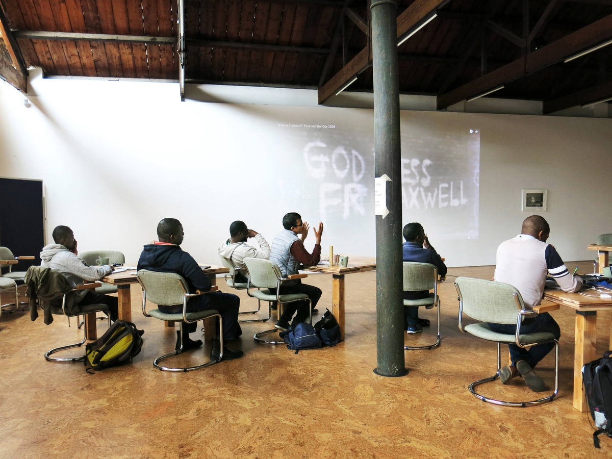 Event photograph from the 2018 rendition of the City Research Studio exchange with the African Centre for Cities. At the front, participants are seated at wooden tables arranged along the room. At the back, a wall projected still image from Terence Davies’ documentary ‘Of Time and the City’.
