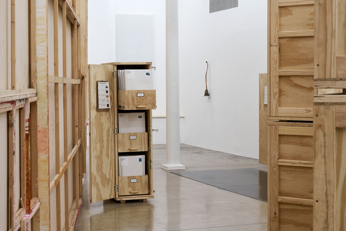 Installation photograph from the 'A Little After This' exhibition in A4 Arts Foundation's gallery. At the back, Lucas Sithole's carved yellowwood sculpture 'Not You!' sitting on a small wall-mounted shelf. On the left, an installation of Penny Siopis' glue, ink and oil 'Maitland' paintings suspended from the ceiling. On the right, collected objects from Penny Siopis' 'Will' work are stored in envelopes and wooden crates.
