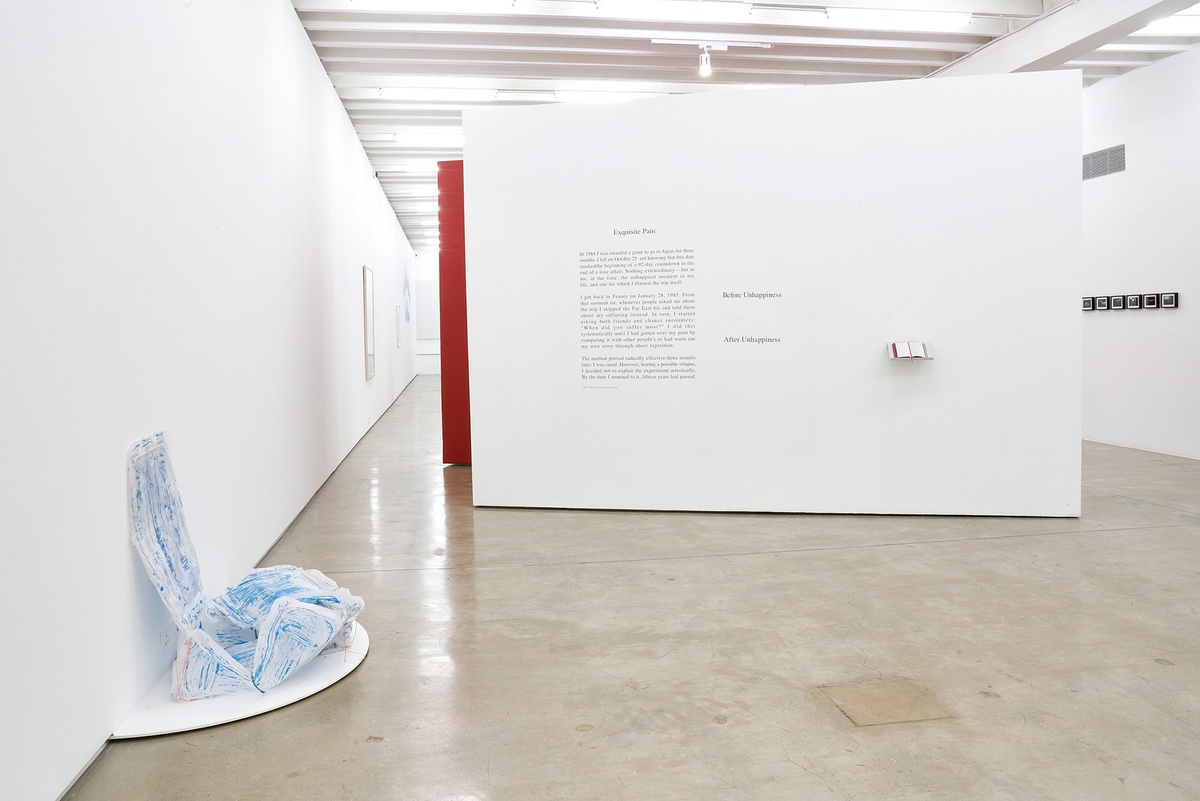 Installation photograph of the You to Me, Me to You exhibition. On the left, Thato Makatu’s sculptural butcher-paper work ‘home is…’ sits on a white semi-circular platform attached to the base of a white gallery wall. At the back, Sophie Calle’s ‘Exquisite Pain’ is arranged on a white moveable gallery wall, with a vinyl wall text on the left and a book on a wall-mounted shelf on the right.

