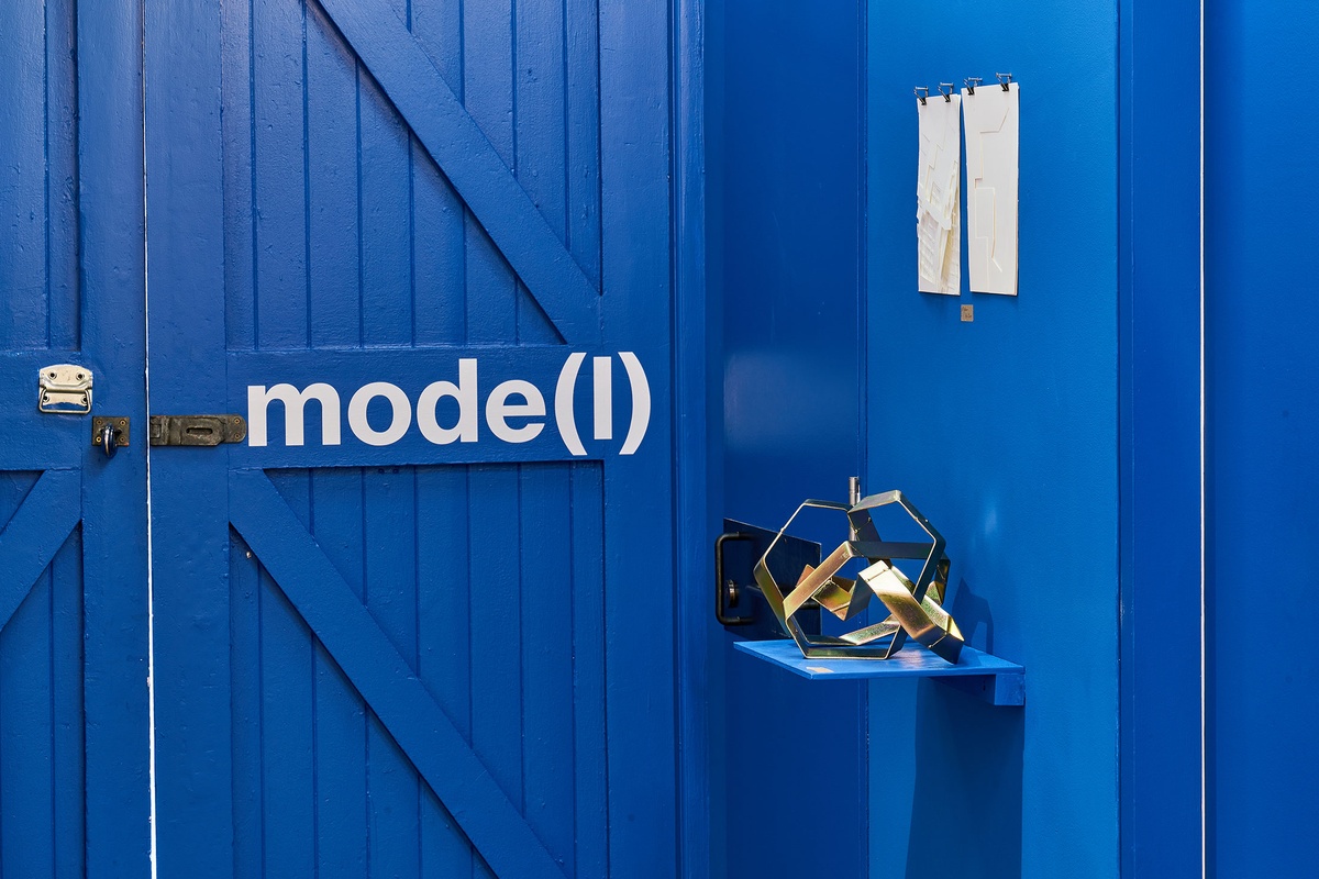 Installation photograph from the ‘mode(l)’ exhibition in A4’s Goods project space. On the left, the exhibition title sits on the wall in white vinyl. On the right, Kyle Morland’s cadmium plated mild steel sculpture ‘40/3 v9.0 for 200/10 4.8’ sits on a blue shelf, with Meghan Ho-Tong’s untitled paper sculpture mounted on the wall above it.
