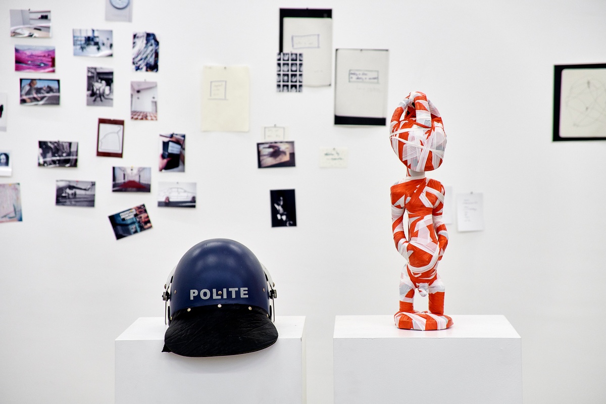 Installation photograph from the 2018 rendition of ‘Parallel Play’ in A4’s Gallery. At the back, photocopies and photographic prints line the gallery wall. On the left, a helmet from Christian Nerf’s ‘Polite Force’ project sits on a white plinth. On the right, Kendall Geers’ sculpture ‘Twilight of the Idols’ sits on a white plinth.
