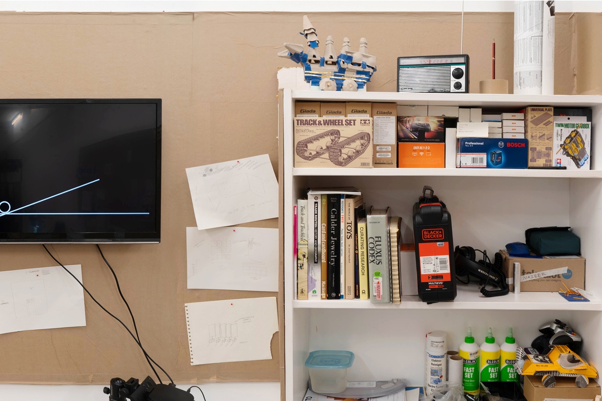 Installation photograph from Mitchell Gilbert Messina's residency in A4 Art Foundation. On the left, a wall-mounted strip of cardboard hosts pinned research notes and a wall-mounted screen. On the right, a bookshelf holds books, electronic components and tools.
