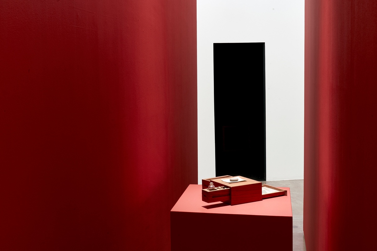 Installation photograph of the You to Me, Me to You exhibition. At the front, Miguel Cinta Robles’ cedarwood box installation ‘Writing device to craft love letters’ sits on a red plinth in between two red moveable gallery walls. At the back, the entrance to the video room is visible.
