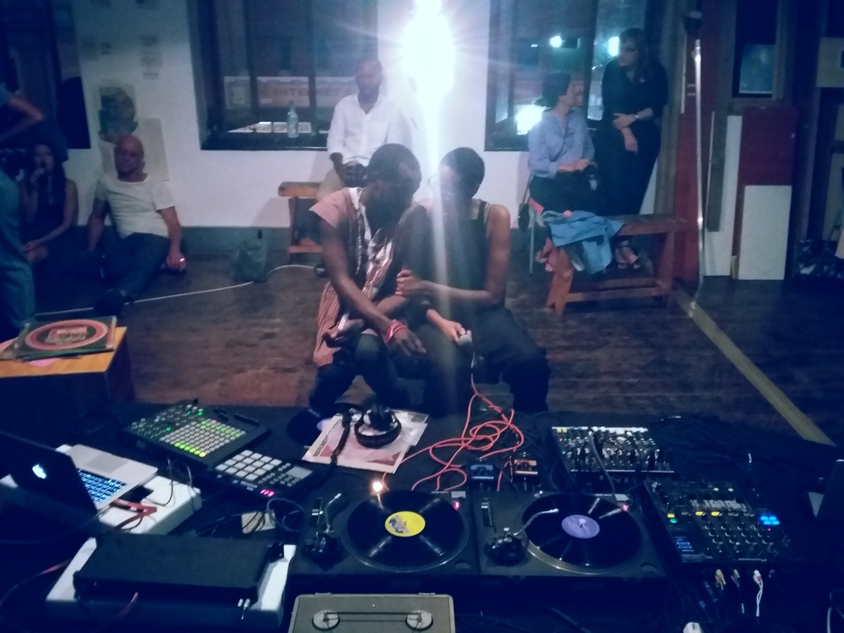 Event photograph from the offsite exchange ‘Pungwe Nights: Listening to Listening’ that shows two participants seated at a table laden with audio equipment, laptops and vinyl records.
