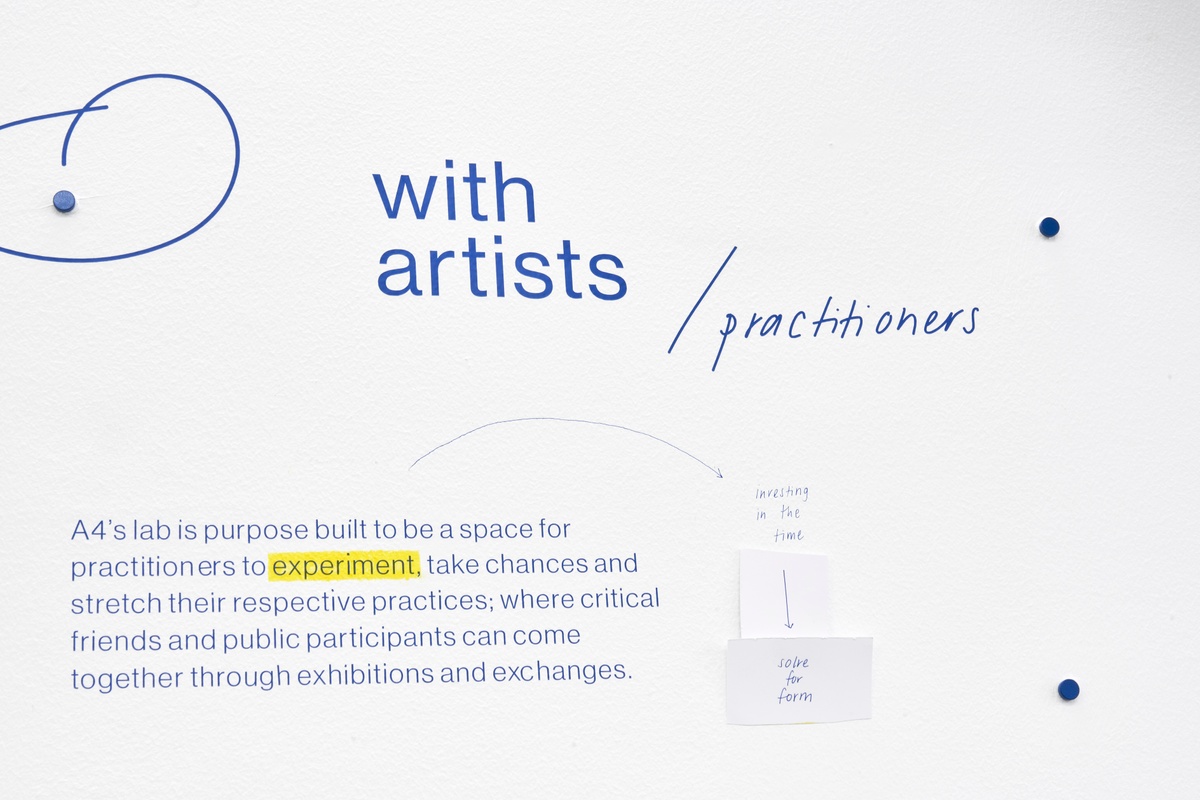 Installation photograph of the A4 About Wall shows the phrase 'with artists/practitioners' described on a white wall in blue. Below, a short explainer text in blue describes A4's relationship to practitioners and public participants.
