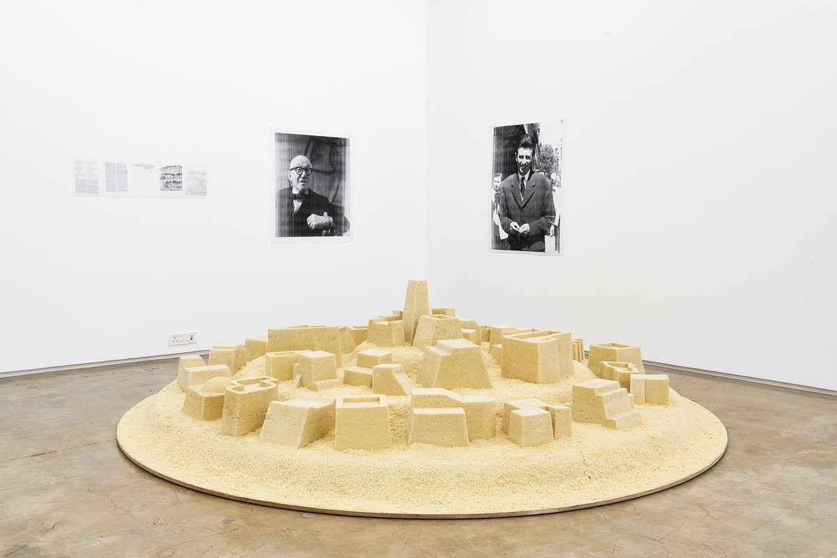 Installation photograph from The Future is Behind Us exhibition in A4’s Gallery. At the front, Kader Attia’s couscous sculpture ‘Untitled (Ghardaïa)’ resembles a town. At the back, photocopied portraits of Le Corbusier and Fernand Pouillon are mounted on the white gallery walls, accompanied by a photocopied UNESCO certificate.
