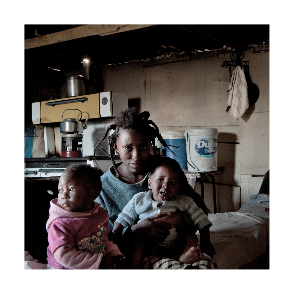 Photograph from Jabulani Dhlamini’s residency on A4’s top floor. At the back, an indoor kitchen area. At the front, a seated individual sits with two children.
