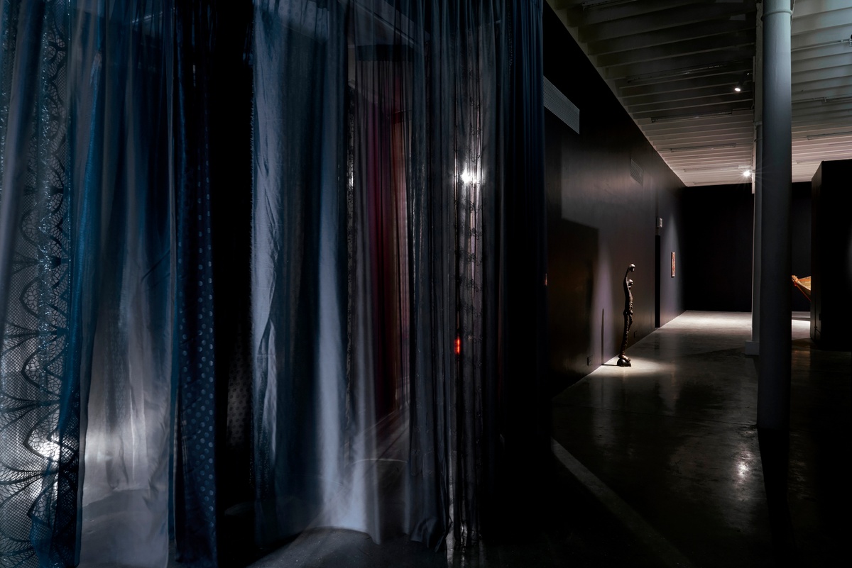 Installation photograph from the ‘Risk’ exhibition in A4’s Gallery. On the left, Igshaan Adams’ untitled fabric installation is suspended from the ceiling. On the right, Lucas Sithole’s bronze sculpture ‘Mother and Child’ stands in front of a wall.
