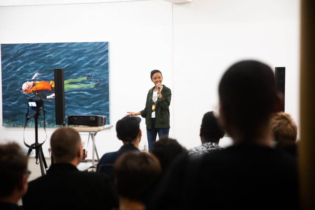 Event photograph from the “Bonolo Kavula presents ‘Art Times’” event in A4’s Library. At the front, attendees are seated in chairs. At the back, Bonolo Kavula performs with a handheld microphone, with Jake Aikman’s oil painting ‘Untitled (portrait of Christian Nerf)’ mounted on the wall behind them.
