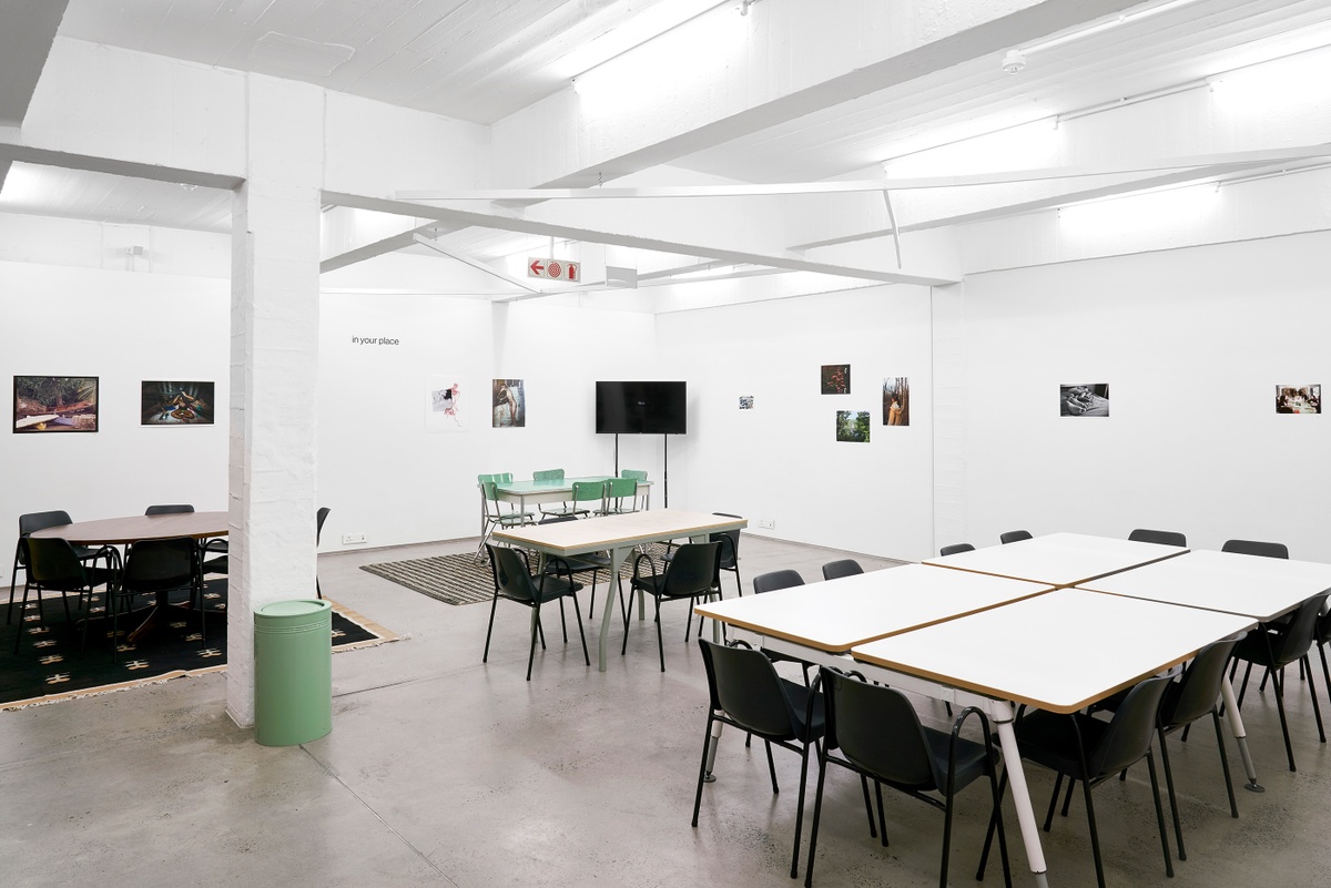 Installation photograph from the ‘In Your Place’ exhibition on A4’s ground floor. At the back, various artworks line the wall. At the front, seating areas with tables and chairs are scattered around the space.
