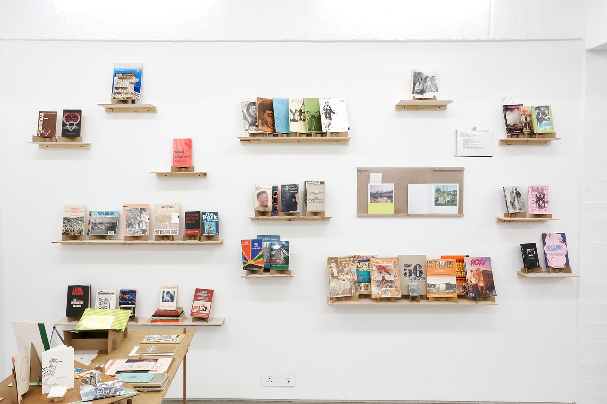 Installation photograph from the Papertrails exhibition in A4’s Reading Room. On the left, a custom wooden table with various printed matter. At the back, various wall-mounted wooden shelves feature printed matter.
