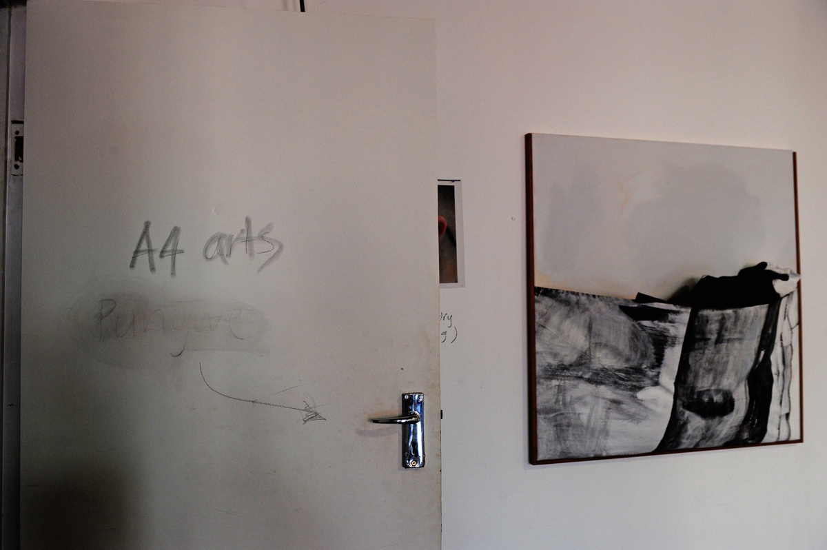 Event photograph from the offsite ‘Office Politics’ exchange. On the left, a white door with the words ‘A4 arts’ and ‘Pungwe’ written in pencil, along with a arrow pointing towards the door handle. On the right, Gerda Scheepers’ acrylic painting ‘Pondoland Pocket’ is mounted on a white wall.
