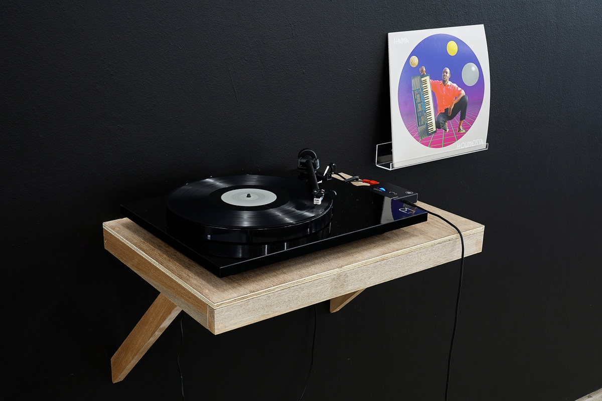 Installation photograph from the ‘Sounding the Void, Imaging the Orchestra V.1’ exhibition in A4’s Gallery that shows a vinyl record player on a wall-mounted shelf with records provided by the Sahel Sounds label.
