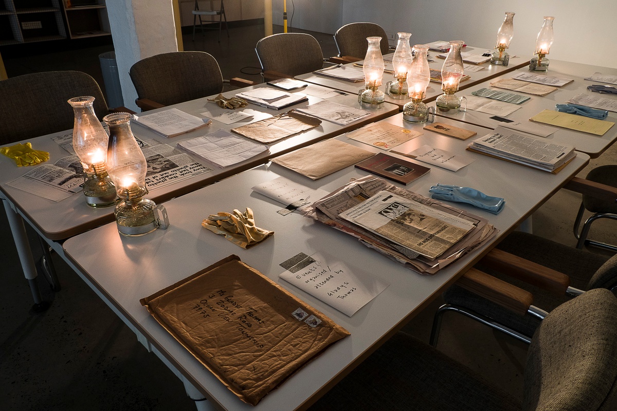 Installation photograph from the ‘Gladiolus’ exhibition on A4’s ground floor that depicts printed matter, oil lamps and archival gloves arranged on four tables.
