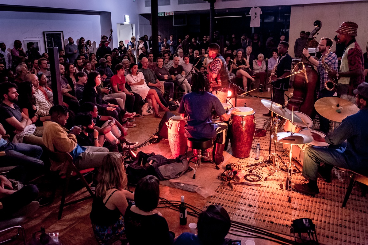 Event photograph from a performance by Shabaka and the Ancestors, with special guest Bra Louis Moholo-Moholo, on A4’s top floor. On the right, the musicians are playing various instruments. On the left, attendees are seated in a curved pattern around the performance area.
