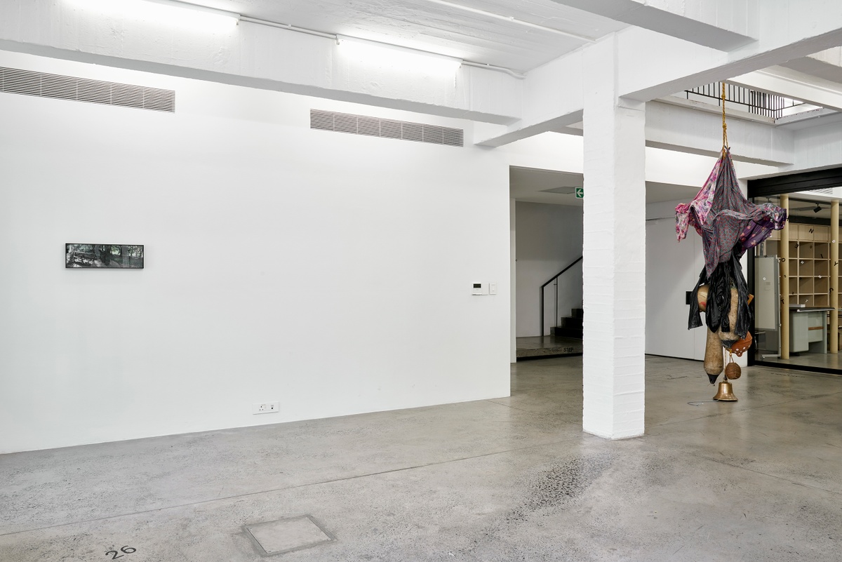 Installation photograph from the ‘without a clear discernible image’ exhibition in A4’s Gallery, showing works hosted on A4’s ground floor. On the left, Kevin Beasley’s photograph ‘Draped’ is mounted on the wall. On the right, Beasley’s untitled resin and garment sculpture is suspended from the gallery floor through an opening in the ceiling.
