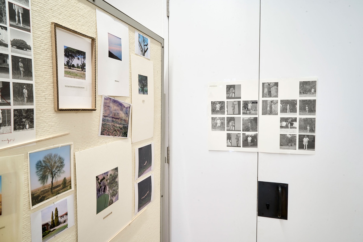 Installation photograph of Michael Tymbios’ studio during his residency in A4 Arts Foundation. On the right, a grid of photographic prints on two sheets are mounted on the doors of a white cupboard. On the left, a freestanding pinboard features multiple text printouts and photographic prints.
