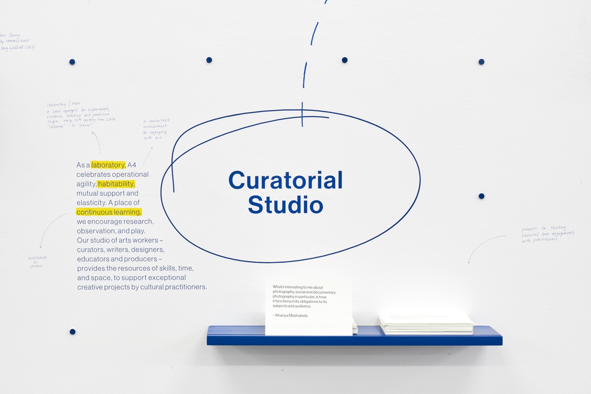Installation photograph of the A4 About Wall shows the phrase 'Curatorial Studio' described and encircled on a white wall in blue. On the left, a short explainer text about A4's curatorial studio. Below, a blue wall-mounted shelf holds printed cards of quotes relating to A4' curatorial studio.
