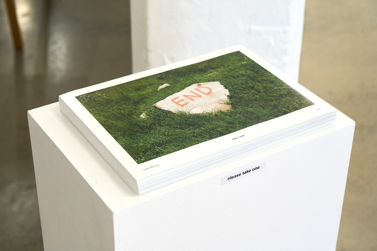 Photograph of a stack of Gabrielle Guy’s lithographic prints ‘END x 1250’, itself a photograph of a rock laying on grass with the word ‘END’ painted on it, sitting on a white plinth.
