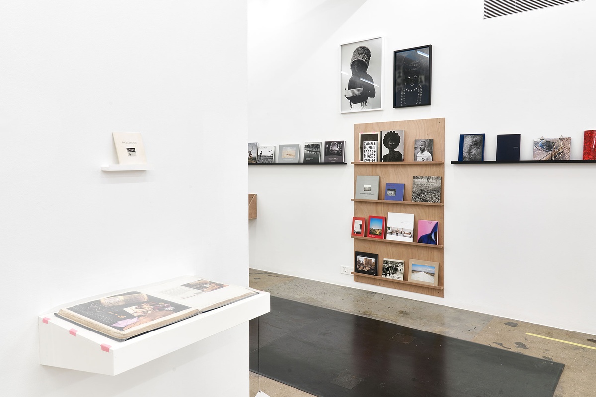 Installation photograph from the Photo Book! Photo-Book! Photobook! exhibition in A4’s Gallery. On the right, shelving units with printed matter from the years 1994 to 2022 line a white wall, along with Zanele Muholi’s photographs ‘Hlanzeka III, Vineyard Hotel, Room 153, Cape Town, 2017’ and ‘Bangizwenkosi, The Sails, Durban’. On the left, shelving units with printed matter mounted on a moveable gallery wall.
