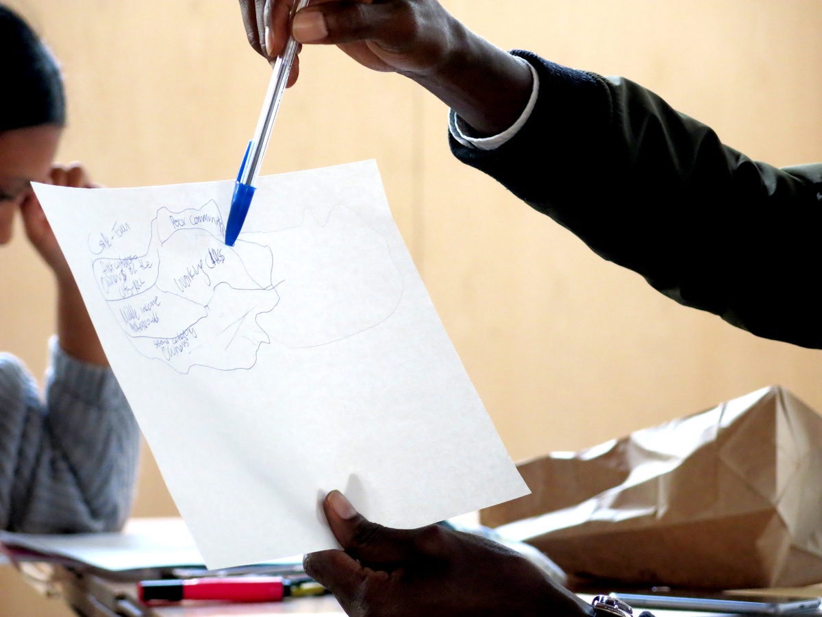 Event photograph from the 2018 rendition of the City Research Studio exchange with the African Centre for Cities. A closeup view of a participant holding a piece of paper with a hand drawn diagram, indicating a specific area with a pen.

