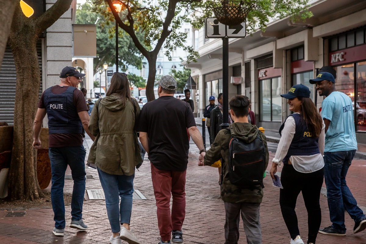 Process photograph from the 2019 rendition of Christian Nerf’s performance piece ‘Polite Force’. Three participants wearing ‘Polite Force’ riot gear escort members of the public along a paved walkway.
