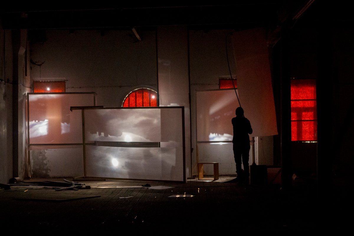 Process photograph from George Mahashe’s residency on A4’s top floor. Four freestanding blank surfaces are arranged around the space so as to catch refracted light from A4’s exterior through Mahashe’s pinhole cameras. On the right, an individual is standing silhouetted in the darkened space.
