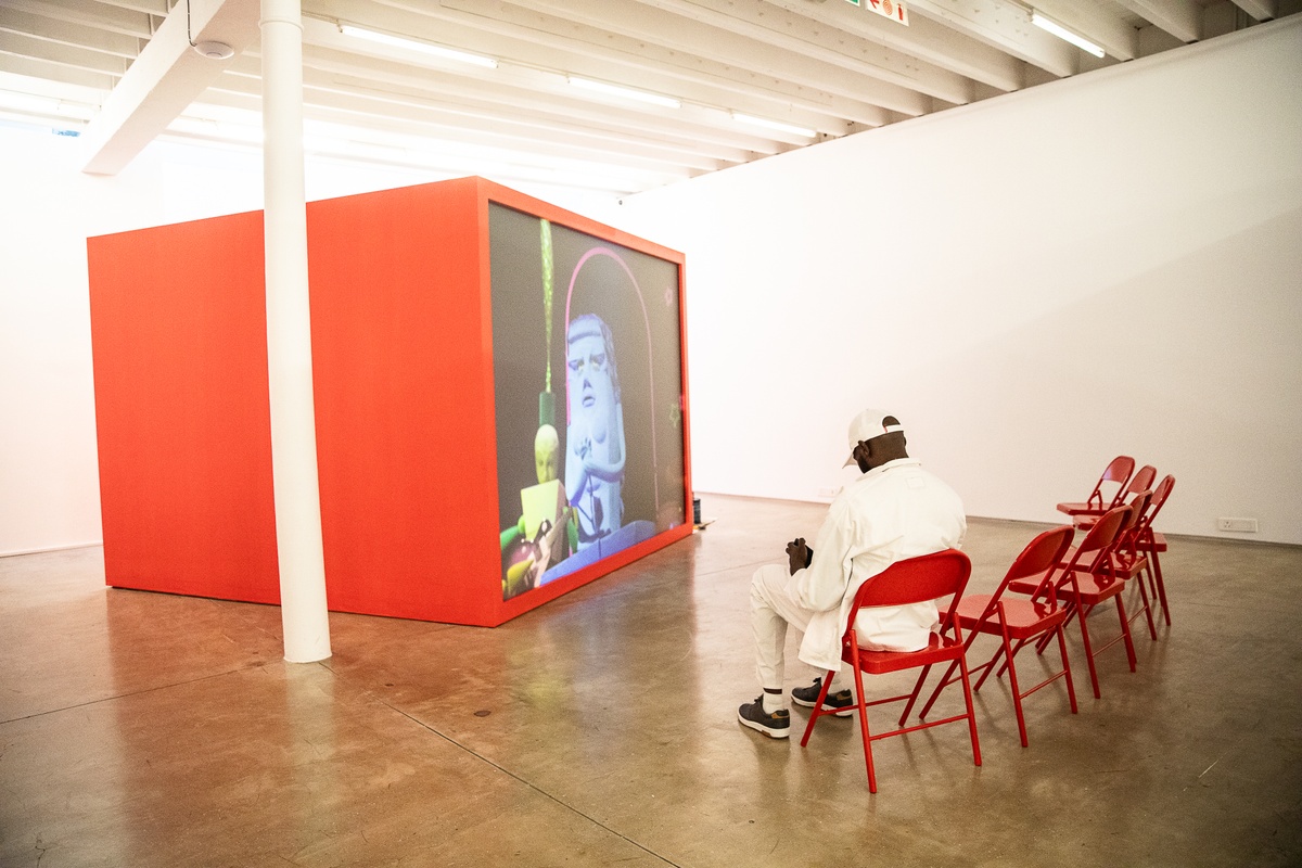 Event photograph from the preview of the 'A Little After This' exhibition in A4 Arts Foundation that shows Alex Da Corte's video installation 'ROY G BIV'. On the left, a large red wooden box with a back-projected screen plays Da Corte's video. On the right, 7 red powder-coated viewing chairs are arranged in an arch. An individual in white overalls sits on one of the chairs.
