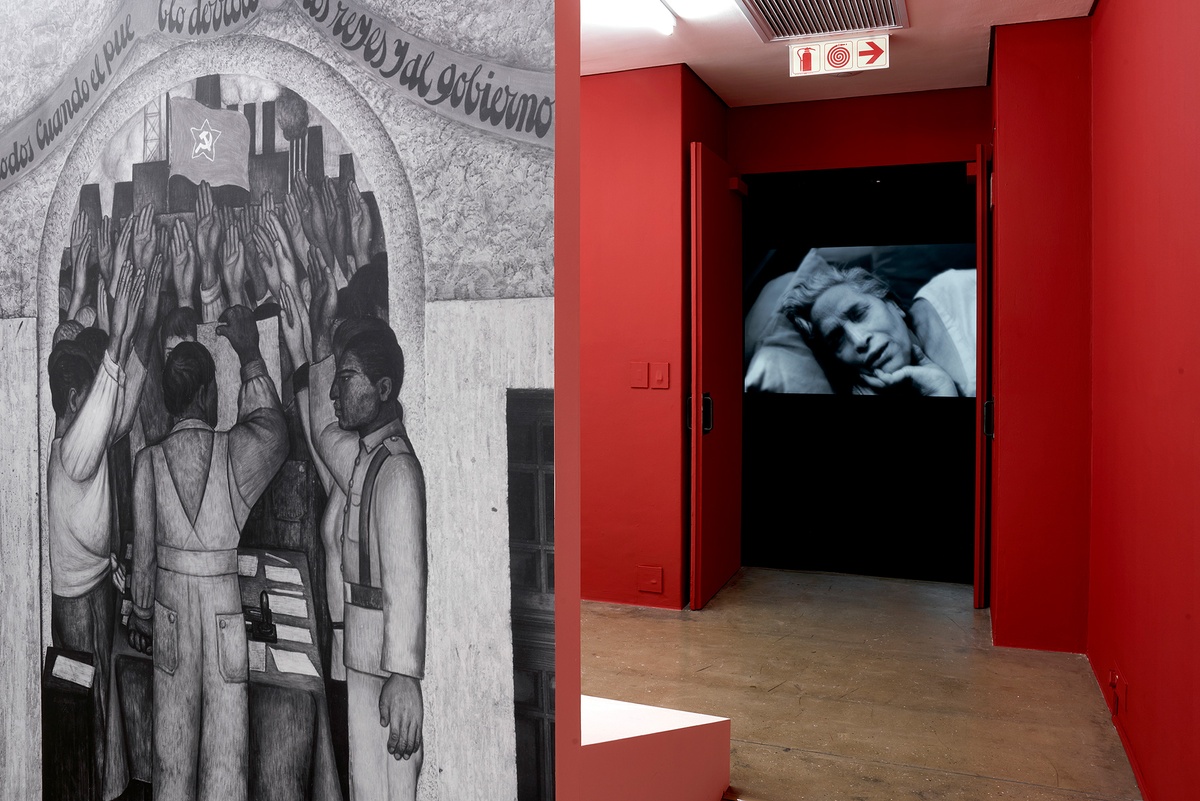 Installation photograph of the You to Me, Me to You exhibition. On the left, Tina Modotti’s photograph ‘La protesta. Corrido de la Revolución.’ is reproduced as wallpaper on a freestanding wall. On the right, Dayanita Singh’s projected video work ‘Mona and Myself’ is partially visible through the video room door.
