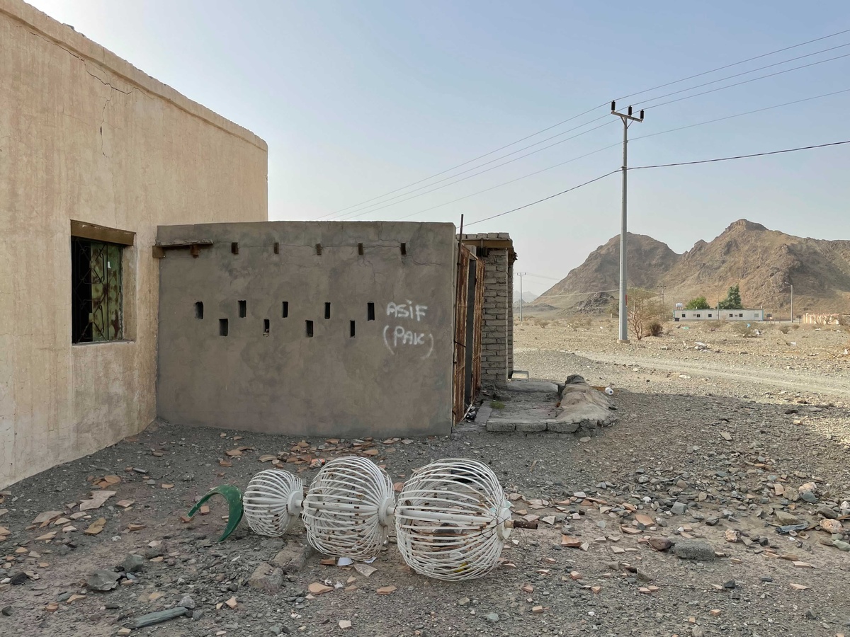 Process photograph from Waiting Upon, Sumayya Vally’s Course of Enquiry at A4, depicting a roadside mosque in Saudi Arabia on the left. At the front, a metal pole with a sickle moon symbol attached lies on the ground. In the middle, an extension of the building is spray painting with the phrases “ASIF” and “(PAK)”. In the back, a telephone line runs next to a dirt road with a mountain in the background.
