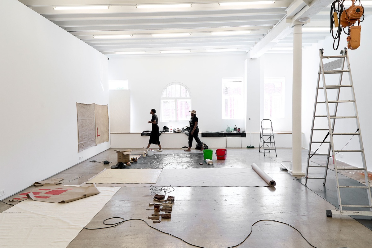 Process photograph of Asemahle Ntlonti’s working space during the 2nd rendition of ‘Parallel Play’ in A4’s Gallery. In the middle, sheets of brown paper and black plastic line the gallery floor, with Ntlonti and curator Kanya Mashabela walking through the space. On the left, two large square paintings are mounted on the wall.
