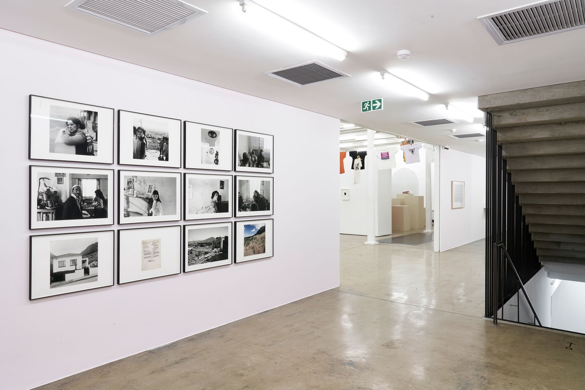 Installation photograph of the Common exhibition. On the left is Sue Williamson’s ‘The Last Supper at Manley Villa,’ a rectangular grid of framed prints hung on a pink wall. On the right is a descending staircase. In the middle is a passage to the main gallery space.
