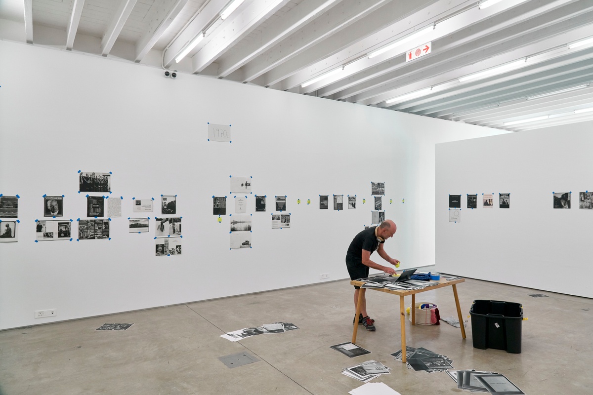 Process photograph from ‘Photobooks’, Sean O’Toole’s Course of Enquiry at A4. At the front, O’Toole is standing at a wooden table laden with photocopies from his photobook collection and his laptop. At the back, photocopies from various photobooks are taped to the walls on the left and right.
