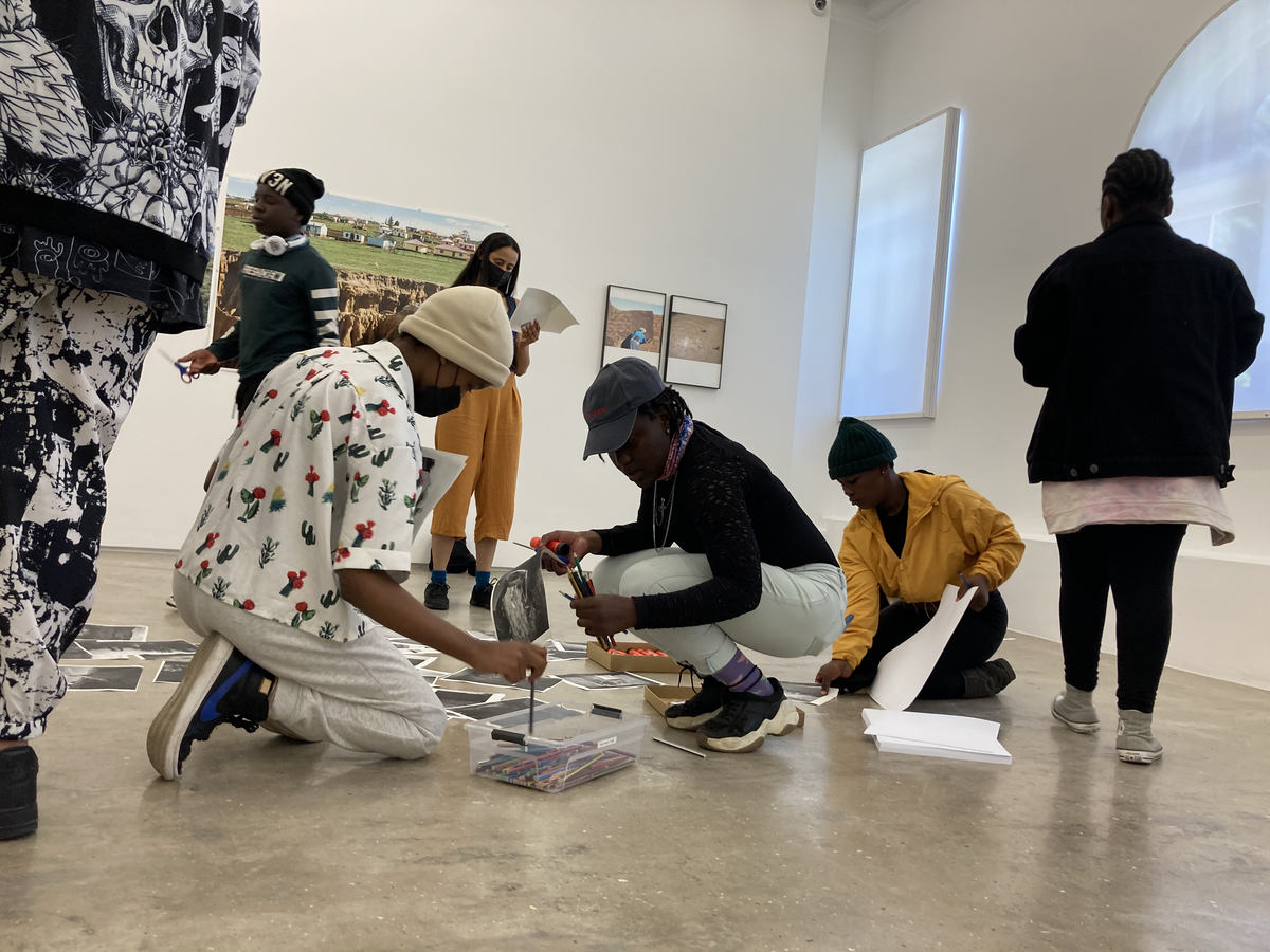 Event photograph from the ‘Lalela x A4' exchange during the ‘Tell It to the Mountains’ exhibition in A4’s Gallery. At the front, Lalela students and facilitators interact with monochrome photocopies of Lidokuhle Sobekwa’s photographs on the gallery floor. At the back, Sobekwa’s photographs ‘Ezindongeni zase Kwezane’, ‘UmamBhele uyatyabeka’ and ‘Trojan river’ are mounted on the gallery wall.
