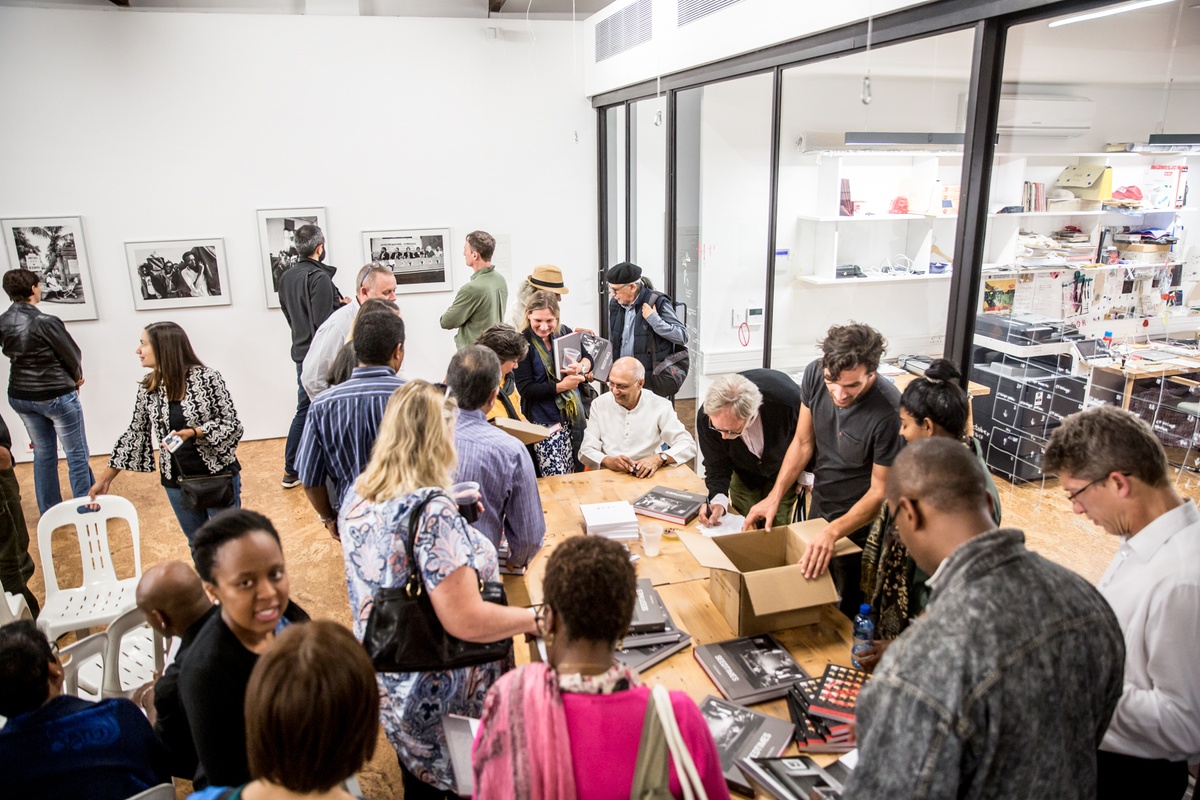 Event photograph from the launch of Omar Badsha’s book ‘Seedtimes’ on A4’s top floor. At the back, attendees look at a row of Badsha’s wall-mounted monochrome photographs. At the front, Badsha is seated at a table with copies of his book surrounded by attendees.
