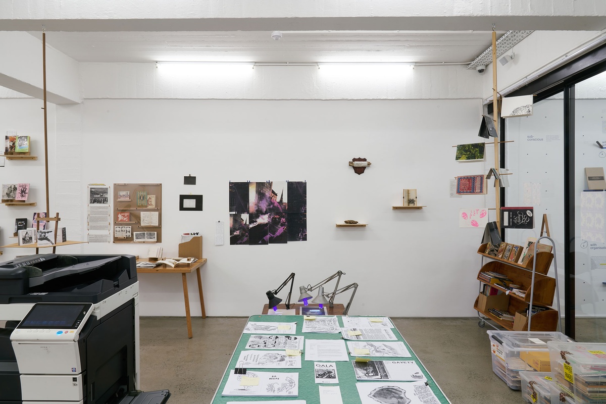 Installation photograph from the Papertrails exhibition in A4’s Reading Room. On the left, a photocopier. At the back, copies of Leonard Shapiro’s untitled photographs of the 1989 Purple Rain protest are pasted onto the wall. On the right, a mobile shelf and plastic crates with printed matter.
