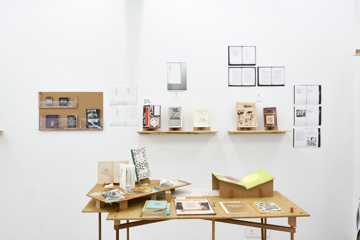Installation photograph from the Papertrails exhibition in A4’s Reading Room. At the front, a custom wooden table with various printed matter. At the back, various wall-mounted wooden shelves feature printed matter.
