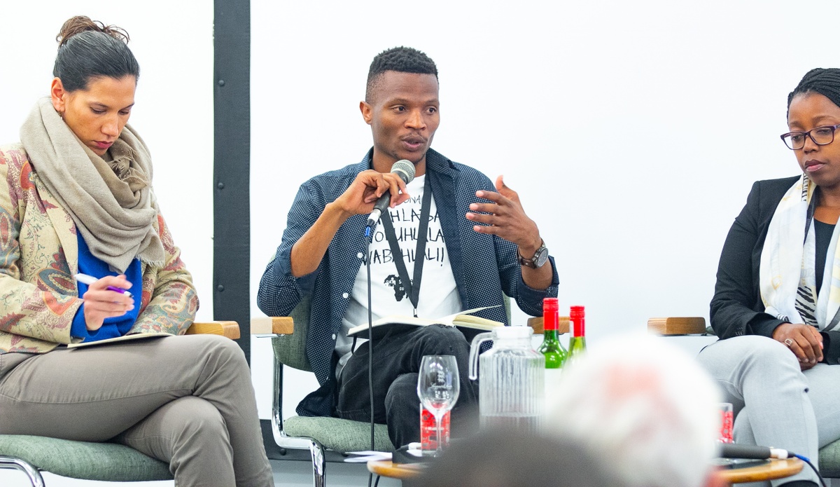 Event photograph from the 2018 rendition of the Open Book festival on A4’s ground floor. At the back, three panelists are seated with microphones.

