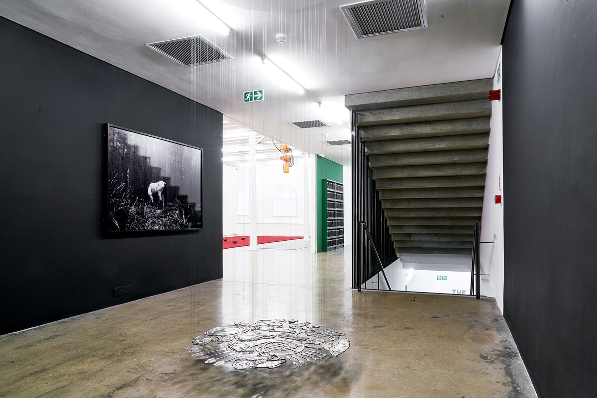 Installation photograph from The Future Is Behind Us exhibition in A4’s Gallery. In the middle, Cornelia Parker’s installation ‘Thirty Pieces of Silver (Exhaled) French Horn’ is suspended from the ceiling. On the left, Santu Mofokeng’s framed monochrome photograph ‘Buddhist retreat near Ixopo’ hangs on a black wall.
