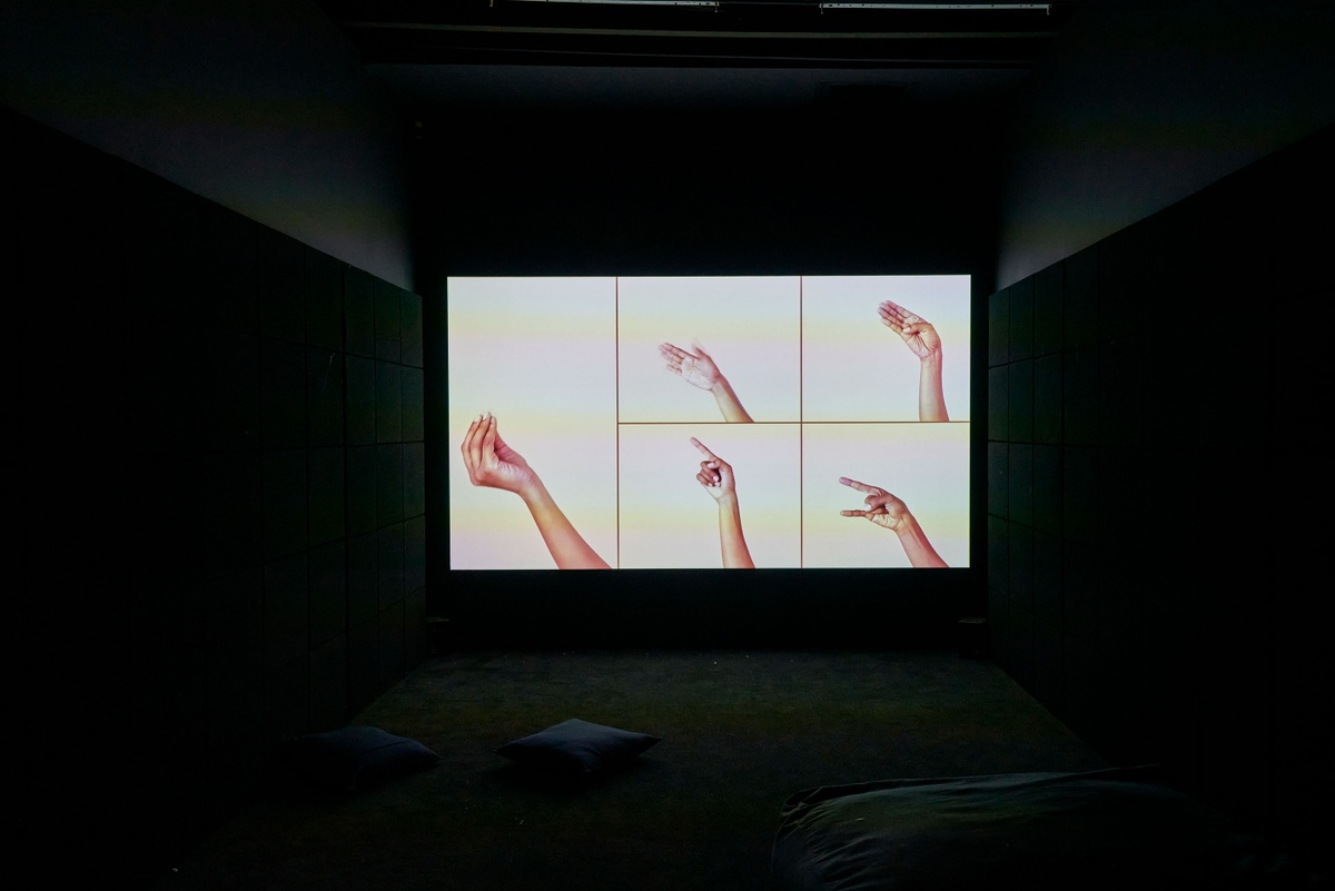 Installation photograph of the Common exhibition. At the back, Lerato Shadi’s video work ‘Mabogo Dinku’ is projected onto a wall in a dark room. The current frame consists of five segments, each featuring a hand making a different gesture against a white background.
