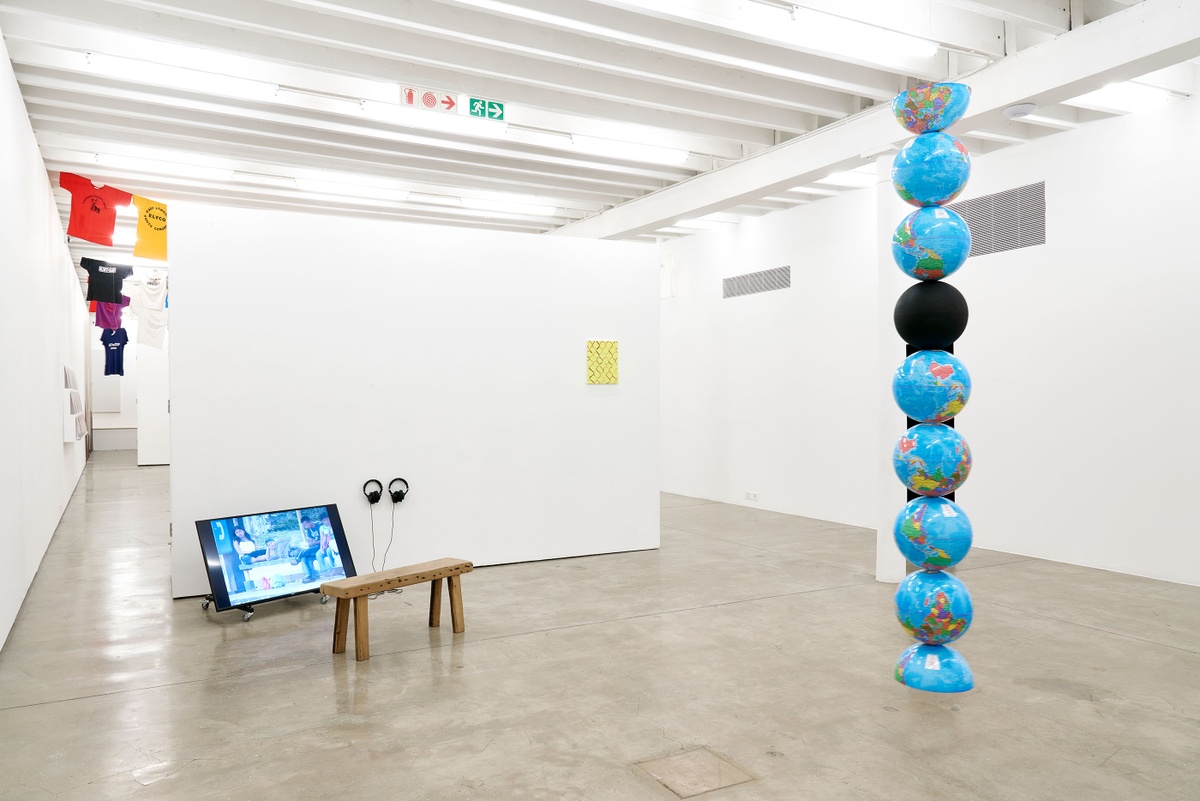 Installation photograph of the Common exhibition. To the right, Nolan Oswald Dennis’ ‘model for an endless column', a column of globe models suspended from the ceiling. One is solid black, the first and last are halved. On the left, a video screen playing Francis Alÿs’ ‘Painting/Retoque' sits on the floor, with earphones and a bench.
