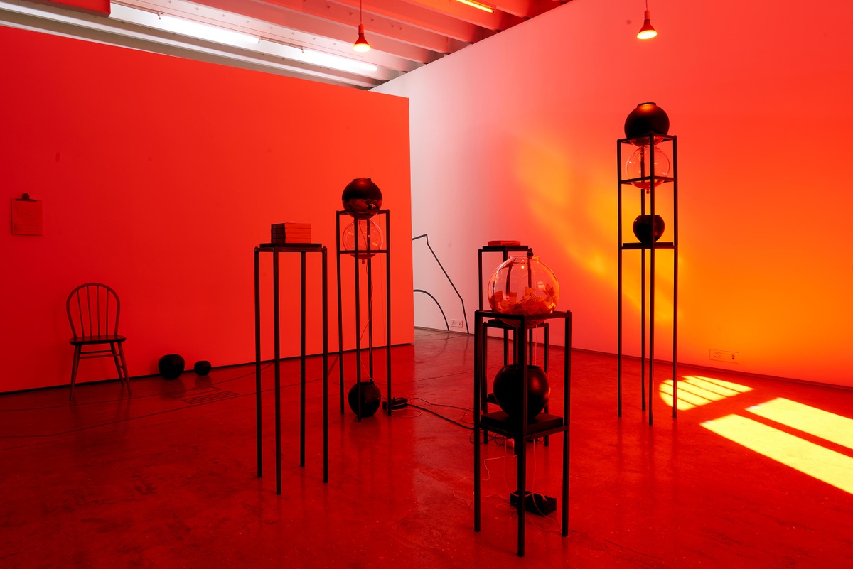 Installation photograph from the Customs exhibition in A4’s Gallery that shows glass globes and steel armatures from Nolan Oswald Dennis’ installation ‘garden for fanon’ sitting on the gallery floor, with the room lit in red.
