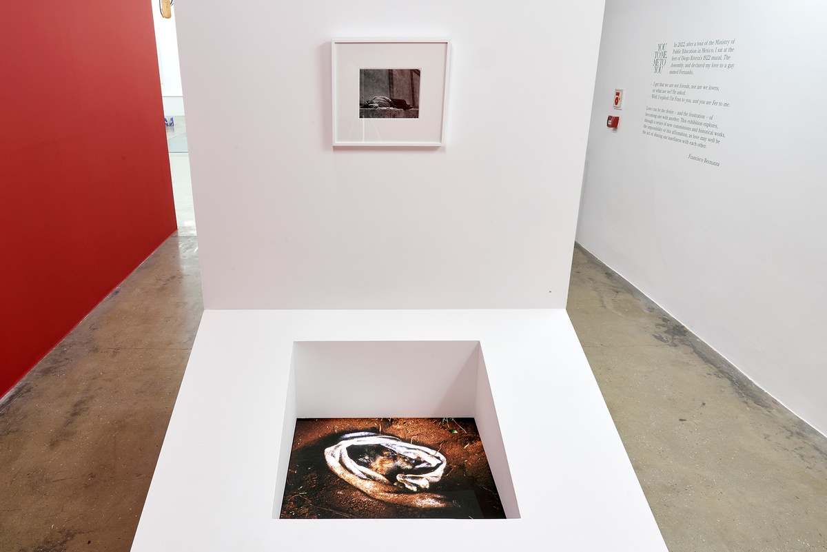 Installation photograph of the You to Me, Me to You exhibition that shows a white freestanding wall with a large slanting support attached to the base. At the top, Manuel Álvarez Bravo’s framed monochrome photograph ‘El Soñador’ is mounted on the wall. At the bottom, Jo Ractliffe’s perspex photographic print ‘Love’s Body’ is embedded in the base, perpendicular to the floor. On the right, the exhibition title and blurb are pasted onto the gallery wall in green vinyl.
