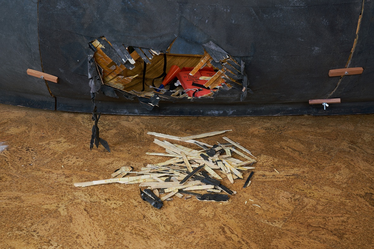 Process photograph from Christian Nerf’s residency at A4 Arts Foundation shows a hole in the side of a rowing boat, with splintered wood lying on the floor in front of it.
